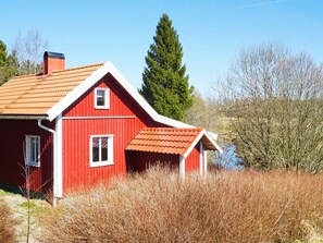 Une maison en bois rouge, avec un toit en pente, entourée d’herbes hautes et d’arbres.