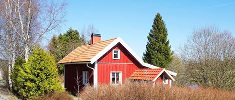 A red wooden house with a sloped roof, surrounded by trees and shrubs.