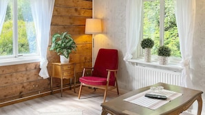 A room with a wooden floor, a red chair, a wooden table with a glass top, a lamp, and a window with curtains. - 4 Person Holiday Home in Hedekas (Hedekas)