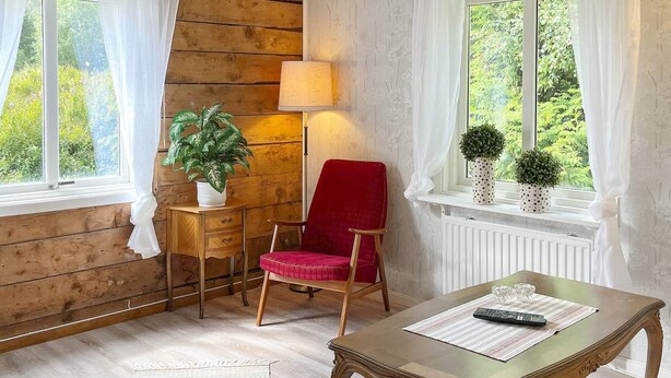 Ein Zimmer mit Holzboden, einem roten Sessel, einem Holztisch mit Glasplatte, einer Lampe und einem Fenster mit Vorhängen.