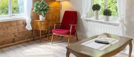 Una habitación con piso de madera, una silla roja, una mesa de madera con cubierta de vidrio, una lámpara y un ventanal con cortinas.
