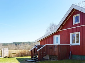 A red building with a white door and windows, a wooden staircase, and a fenced area with a metal gate. - 4 Person Holiday Home in Hedekas (Hedekas)