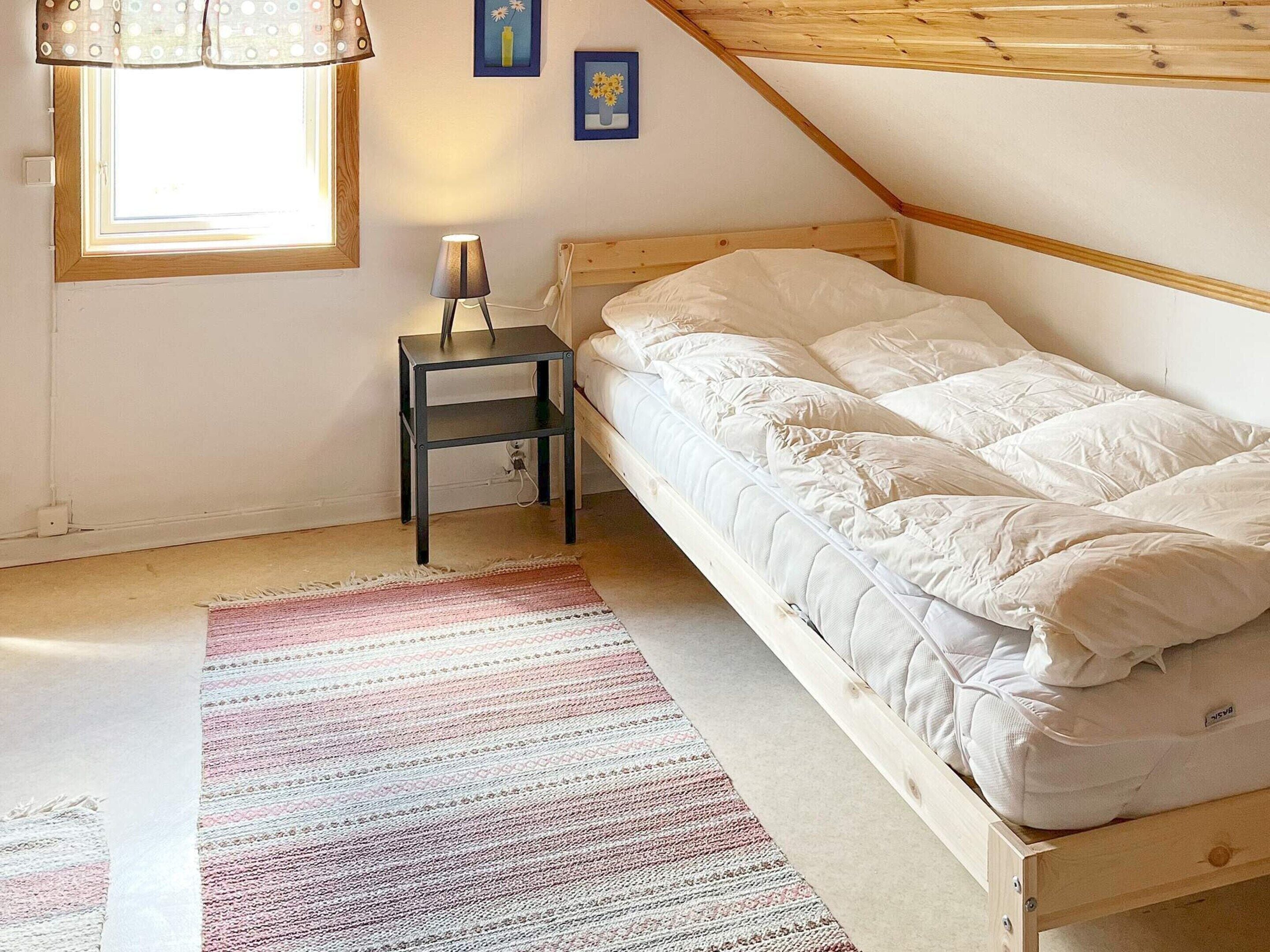A single bed with white bedding in a room with a sloped ceiling, a small bedside table with a lamp, and a colorful striped rug.