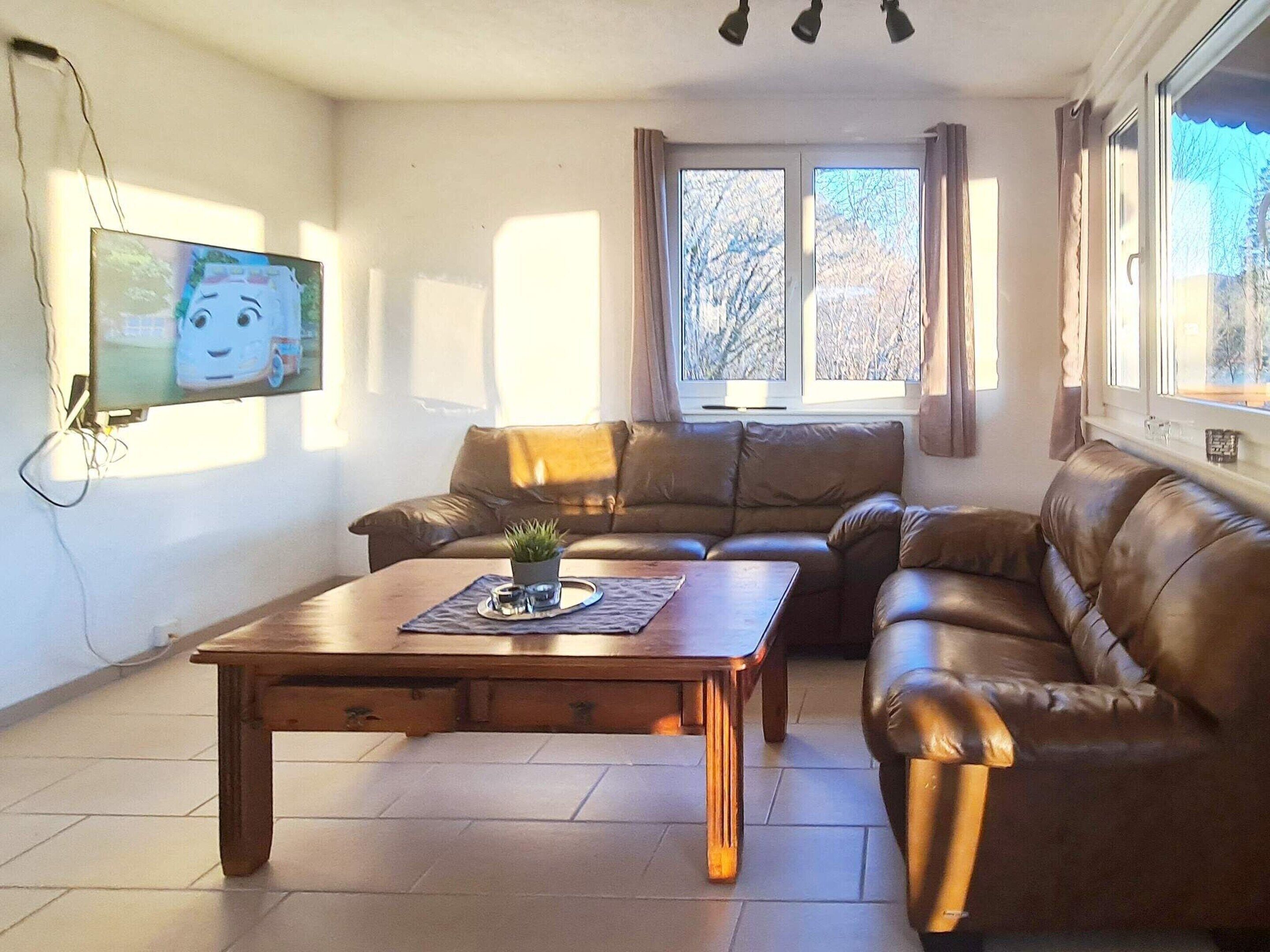 A living room with a brown leather sofa, a wooden coffee table with a plant, and a TV mounted on the wall.