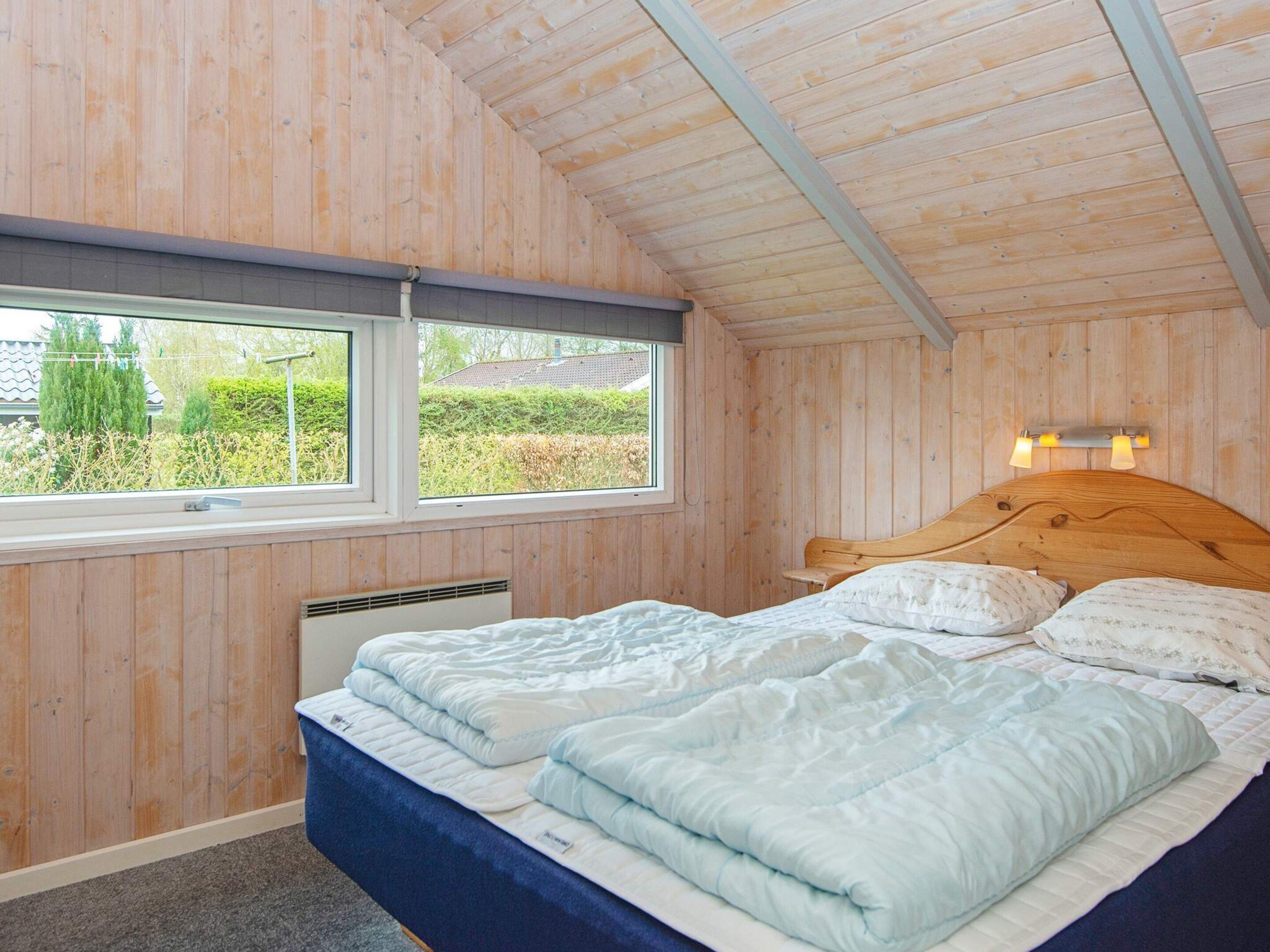 Ein Schlafzimmer mit Holzboden, einem Bett mit weißer Bettwäsche, einem Fenster mit Blick ins Grüne und einer Wandleuchte.