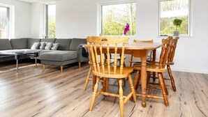 A modern living room with a grey sofa, wooden dining table, and chairs, large windows with a view of greenery, and wooden flooring. - 5 Person Holiday Home in Borkop (Borkop)