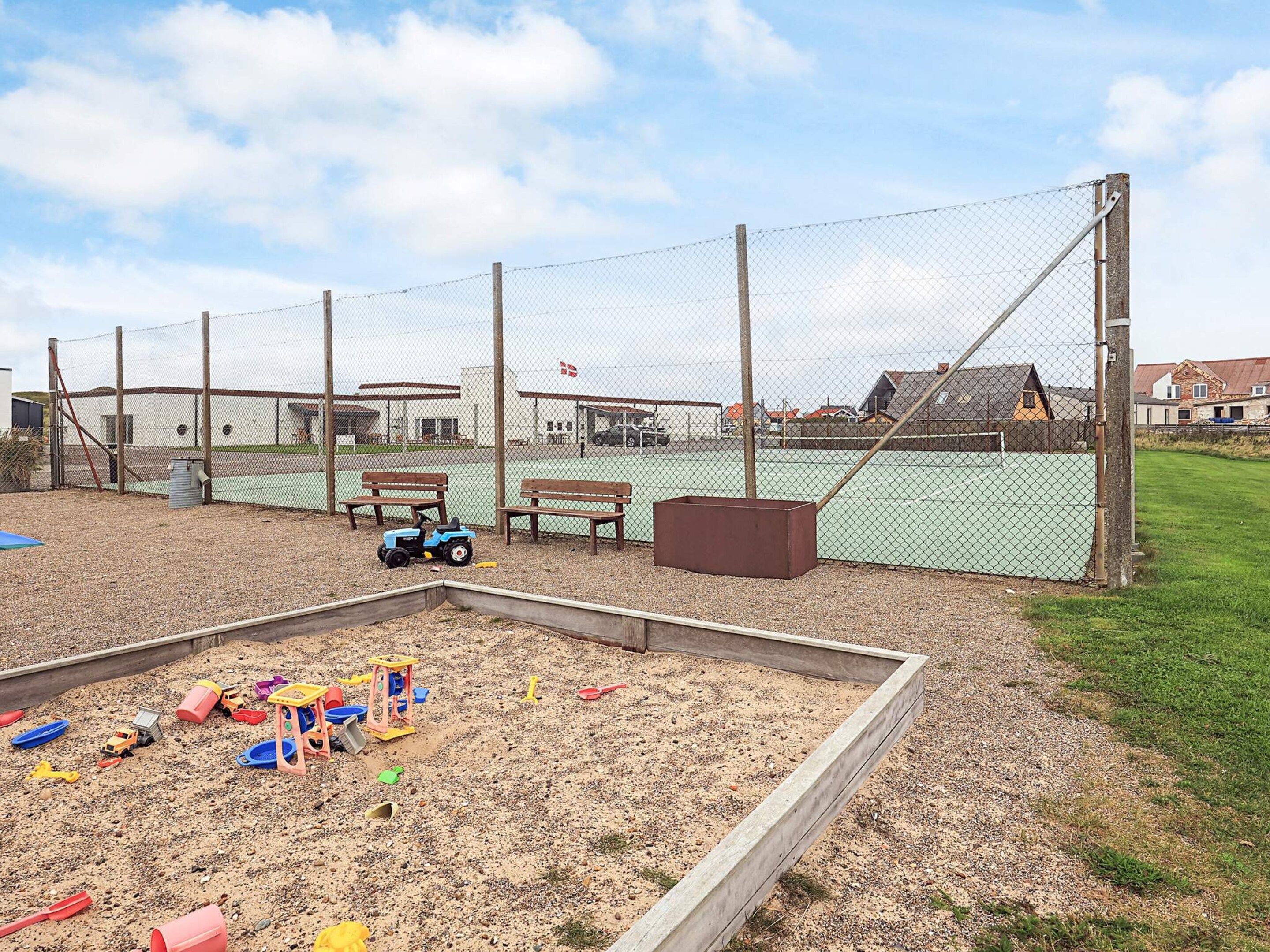 Un área de juegos al aire libre con arena, juguetes, bancos y una cancha de tenis al fondo.