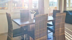 A dining area with a dark wooden table and woven chairs, large windows with curtains, and a view of trees outside.