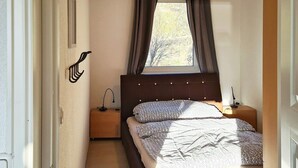 A bedroom with a bed, a nightstand, a lamp, a window with curtains, and a wooden ceiling.