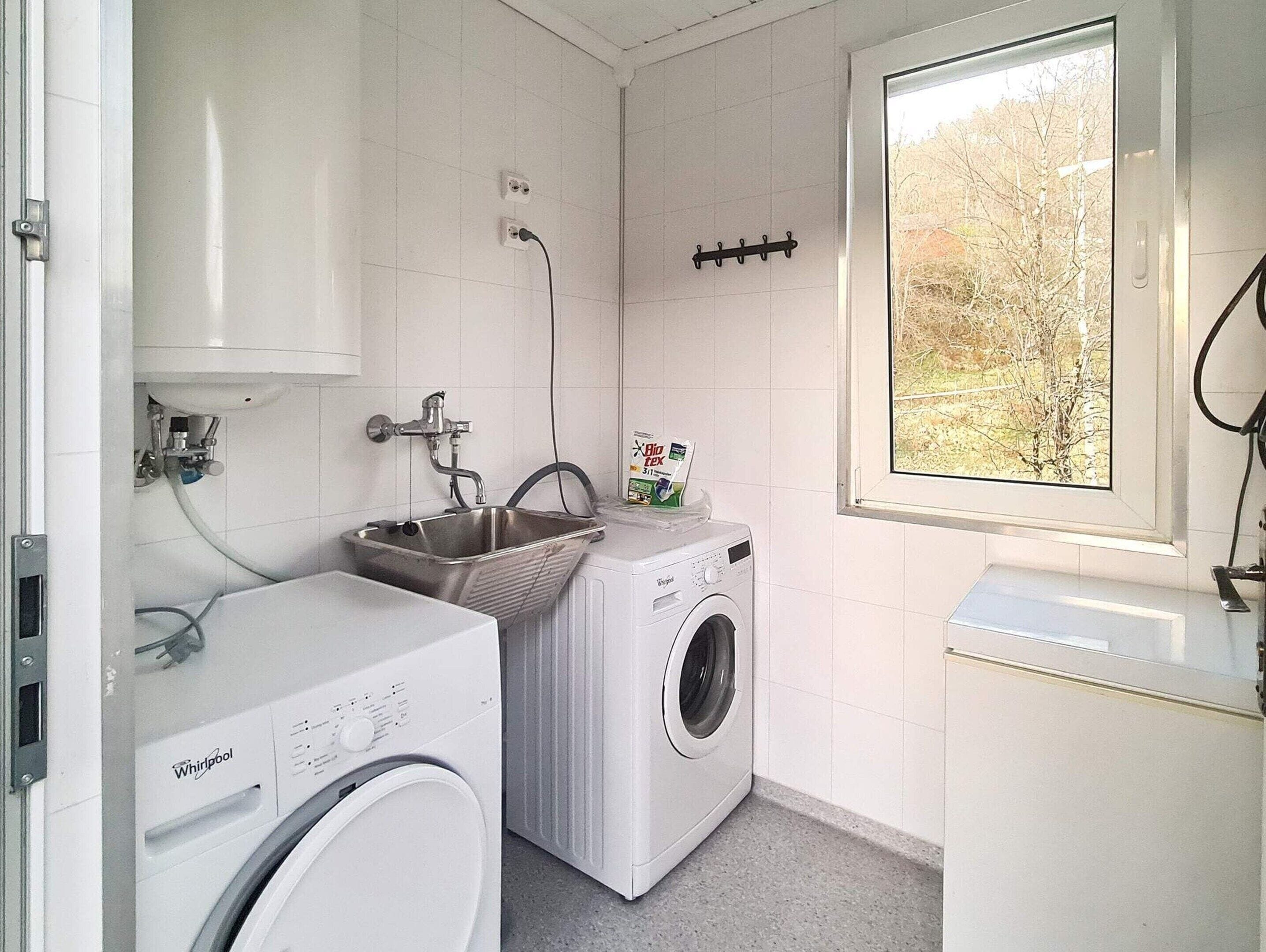 A laundry room with a front-loading washer and dryer, a sink, and a window with a view of trees.