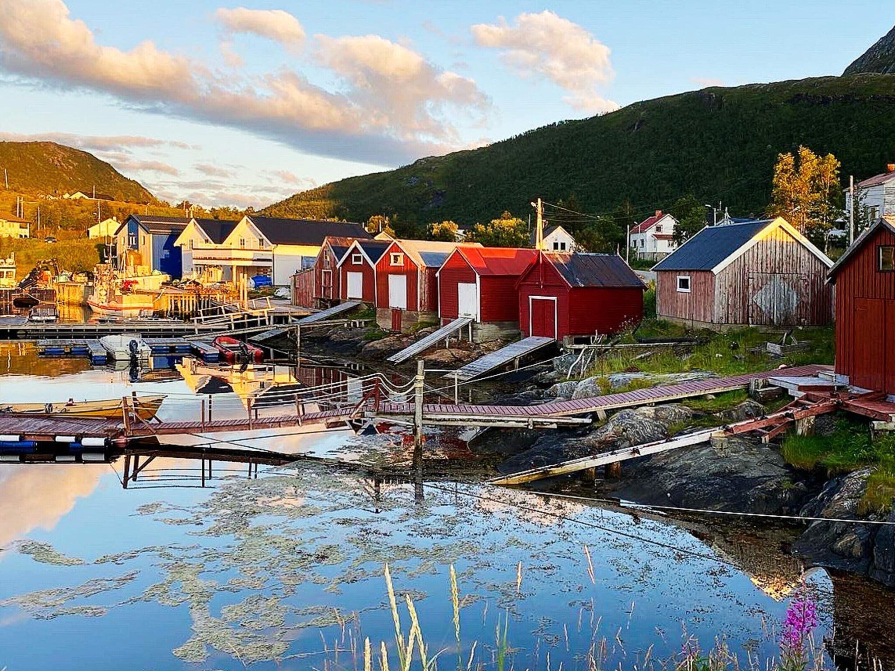 Un pueblo costero con edificios tradicionales de madera roja, un muelle y una masa de agua tranquila que refleja las construcciones.