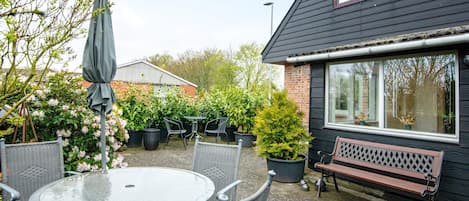 A patio area with a round table and chairs, a bench, potted plants, and a building with a window.