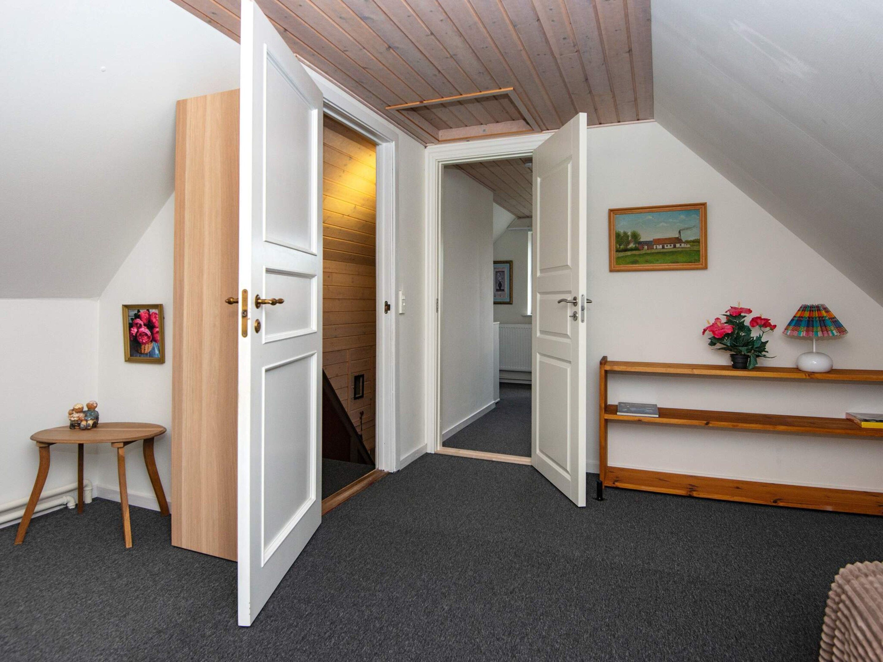 Une pièce avec un plafond en bois, une porte donnant sur un couloir, une petite table avec une plante et une étagère avec une lampe.