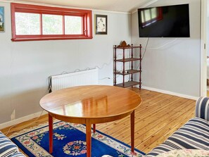 A living room with a wooden table, a blue patterned rug, a striped sofa, a red-framed window, a white radiator, and a mounted television. - 6 Person Holiday Home in Munso (Munsö)
