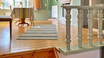 A room with a wooden floor, a striped rug, a dining table with chairs, a staircase with a white balustrade, and a green wall with a mirror and a shelf.