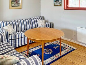 A striped sofa with floral cushions, a round wooden table, and a blue floral rug in a room with wooden flooring and a white radiator. - 6 Person Holiday Home in Munso (Munsö)