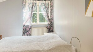 Une chambre à coucher avec un lit, une table de chevet, une lampe et une fenêtre ornée de rideaux à motifs floraux.