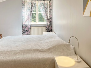 A bedroom with a bed, a bedside table, a lamp, and a window with floral curtains. - 6 Person Holiday Home in Munso (Munsö)