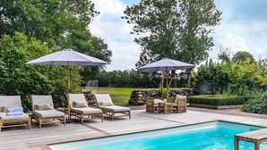 Zona junto a la piscina con sillones reclinables y sombrillas, rodeada de vegetación.