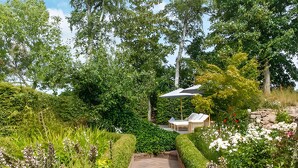 Un sendero de jardín conduce a un pabellón con una mesa y sillas, rodeado de exuberante follaje y plantas florecientes.