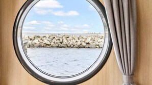 Une fenêtre ronde offrant une vue sur une côte rocheuse et une étendue d’eau calme.