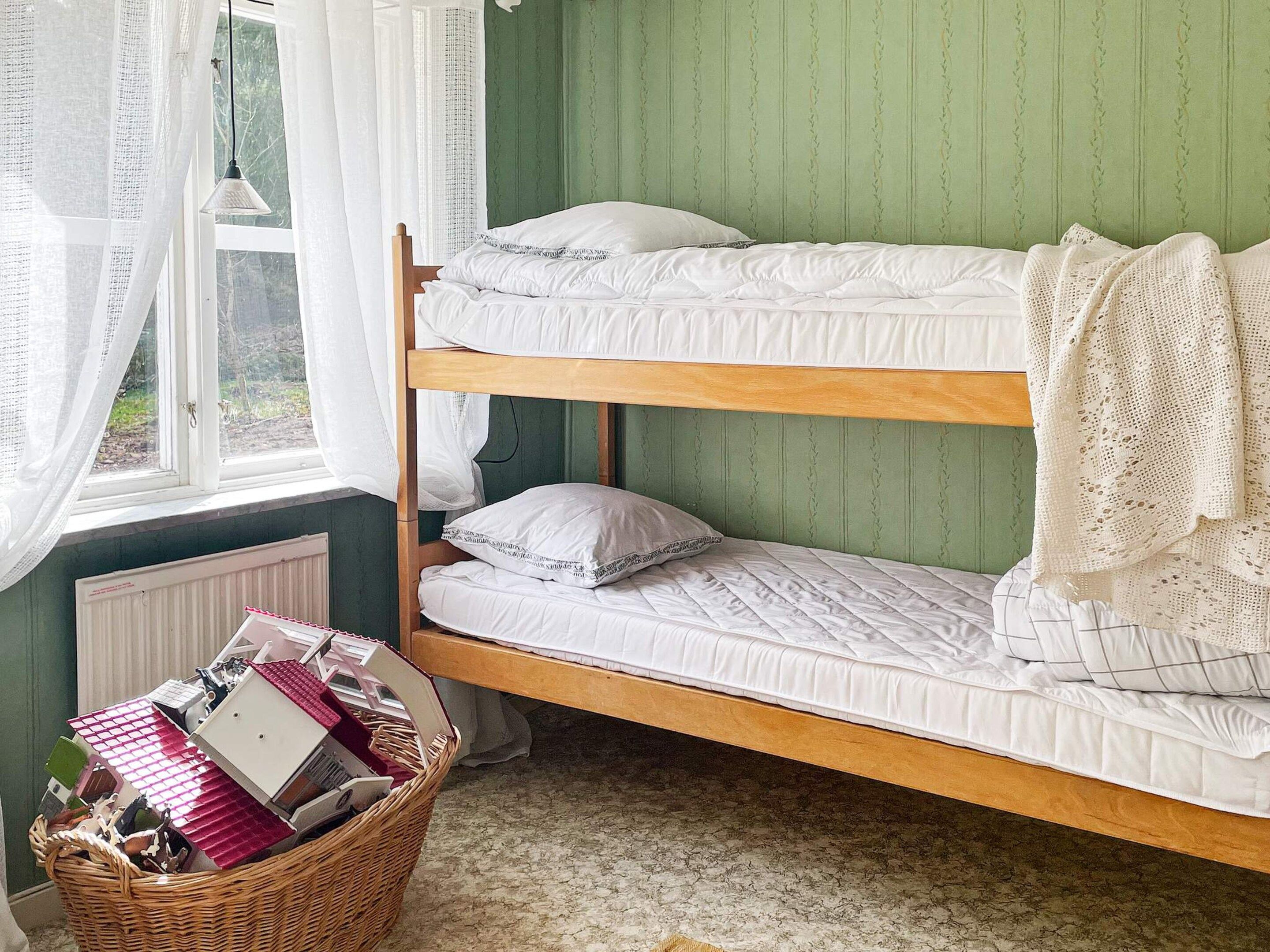A bunk bed with white bedding, a wicker basket with books, and a window with sheer curtains.