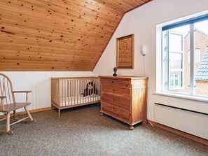 A room with a sloped wooden ceiling, a crib, a wooden dresser, a chair, and a window with a view of a brick building. - 6 Person Holiday Home in Aeroskobing (Aeroskobing)