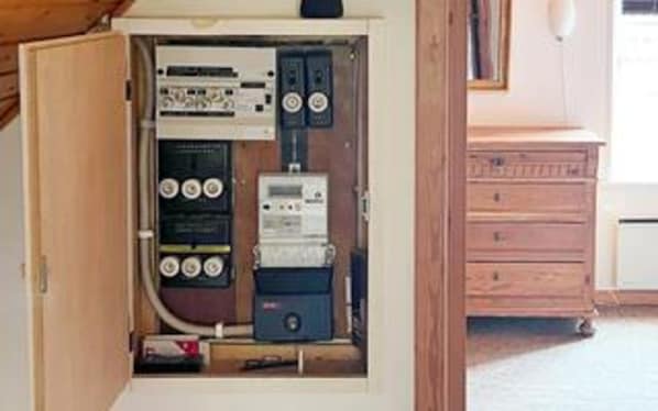 Un tablero eléctrico con varios interruptores y medidores, un armario de madera y un electrodoméstico blanco al fondo.