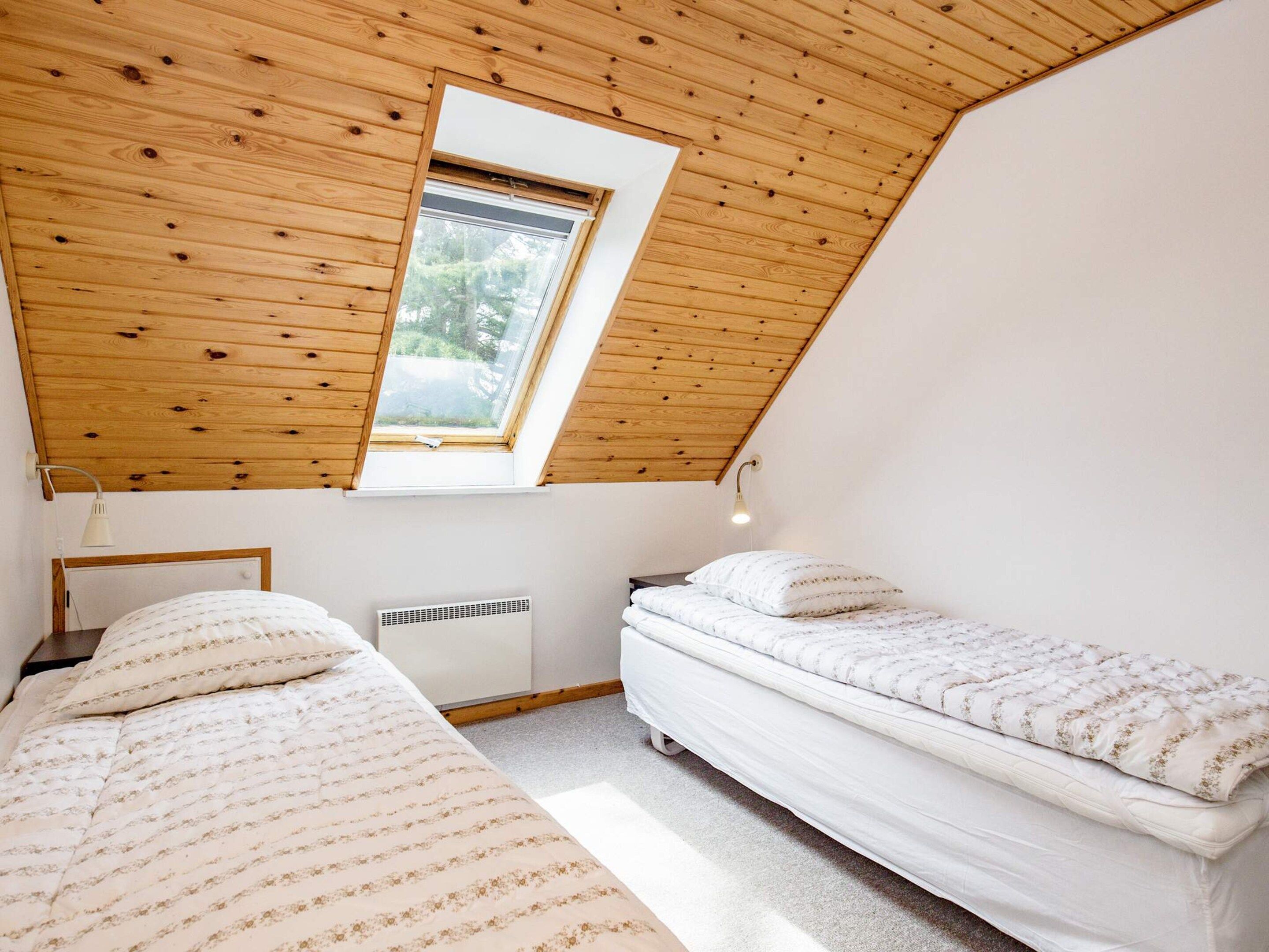 A small, single-room accommodation with a sloped ceiling, a skylight, two beds, a bedside table, and a wall-mounted heater.