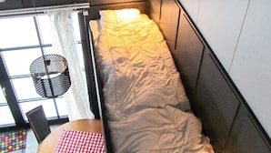 Une petite pièce, de taille modeste, comprenant un lit, une table recouverte d’une nappe à carreaux et une fenêtre avec des rideaux.