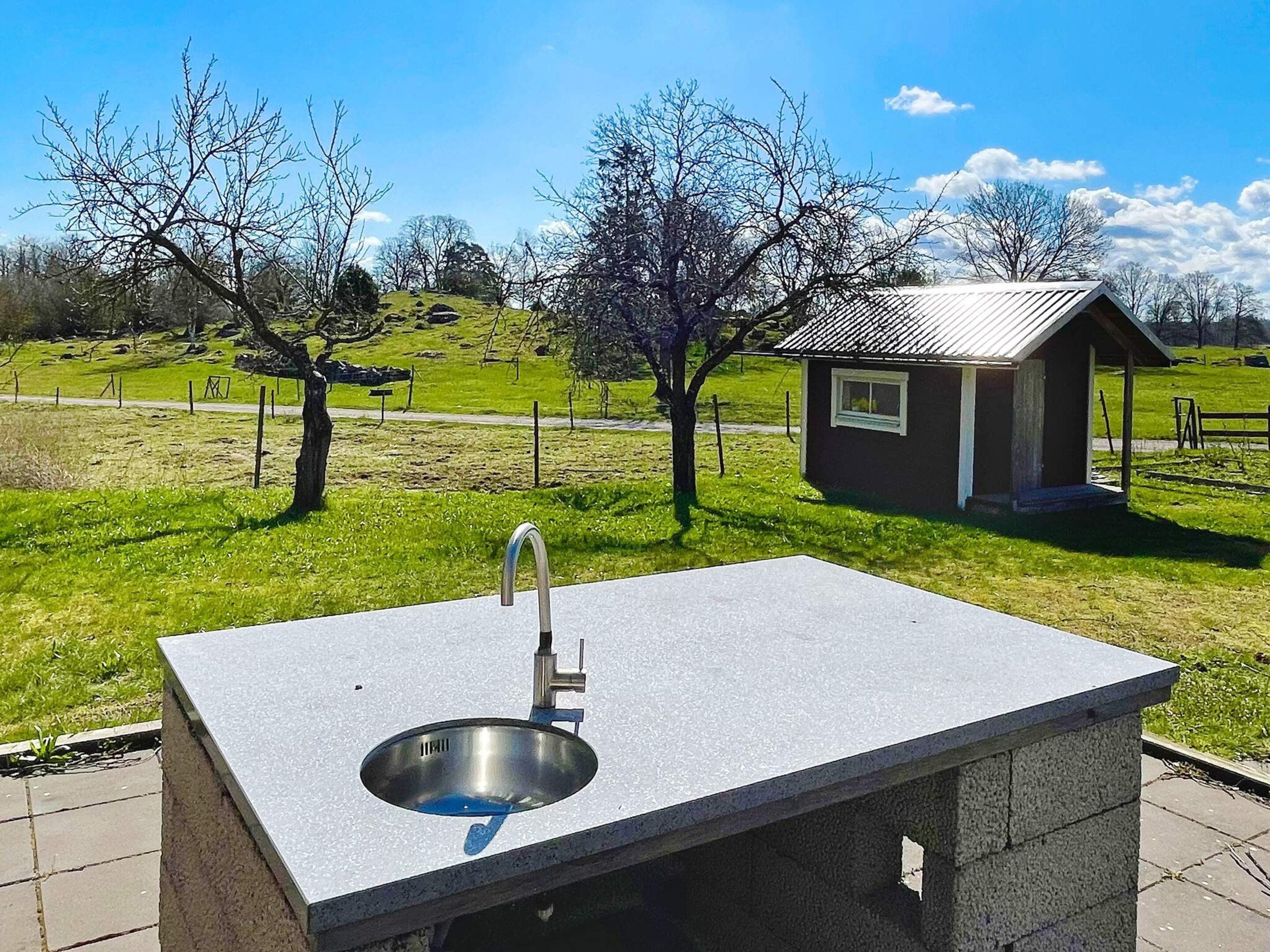 A concrete outdoor sink with a metal basin, a small building with a sloped roof, and a grassy field with trees in the background.