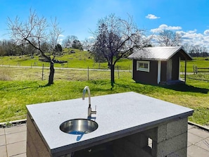 A concrete outdoor sink with a metal basin, a small building with a sloped roof, and a grassy field with trees in the background. - 4 Star Holiday Home in Morlunda (Mörlunda)
