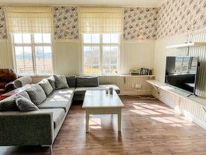 A living room with a sectional sofa, a coffee table, a television, and large windows with patterned wallpaper.