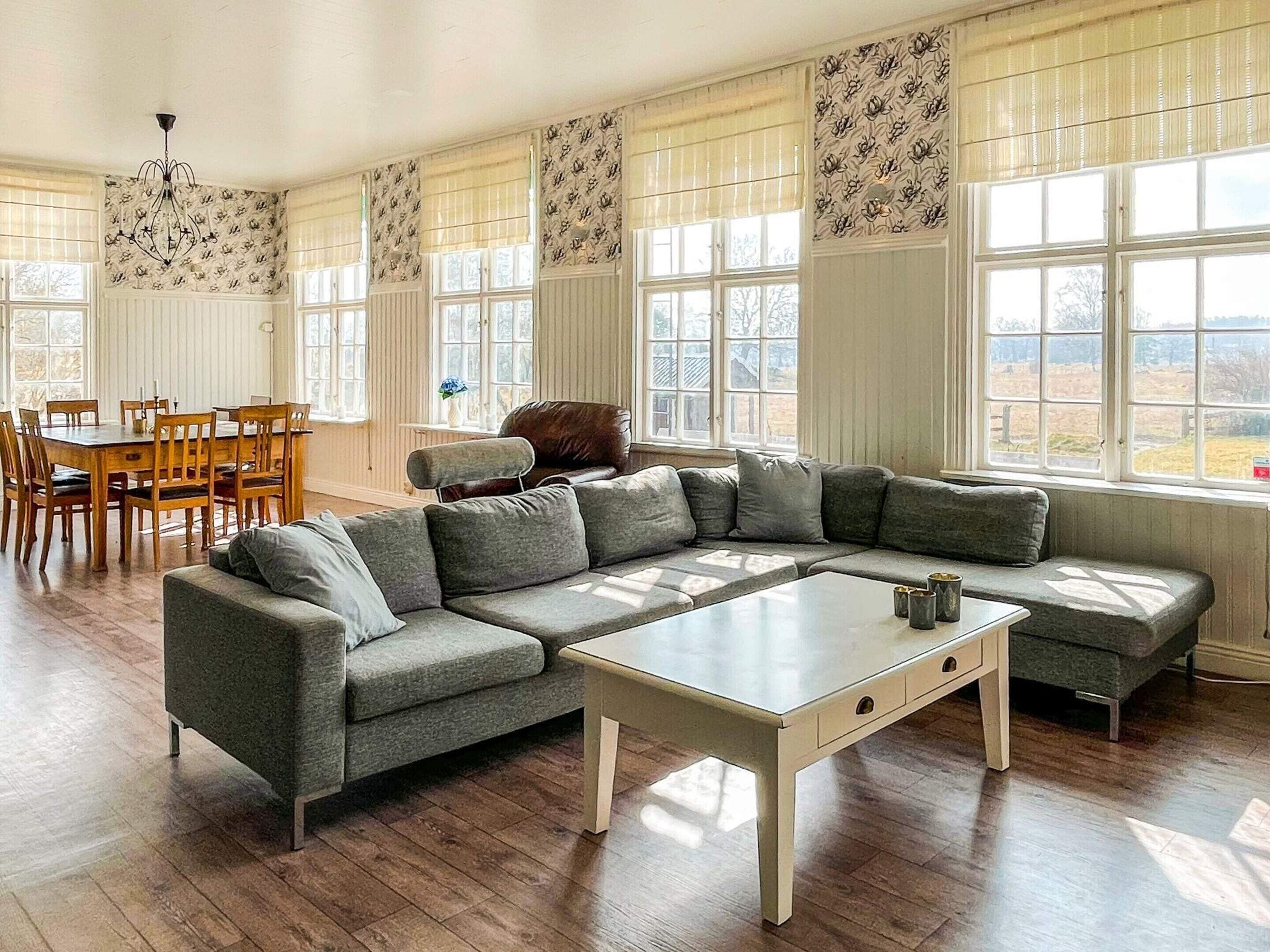 A room with a sectional sofa, a dining table with chairs, and large windows with patterned curtains.