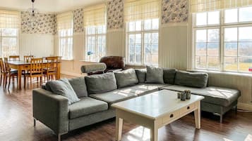 A room with a sectional sofa, a dining table with chairs, and large windows with patterned curtains.