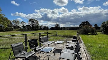 An outdoor seating area with metal chairs and tables, surrounded by a wooden deck and a grassy field, under a partly cloudy sky.