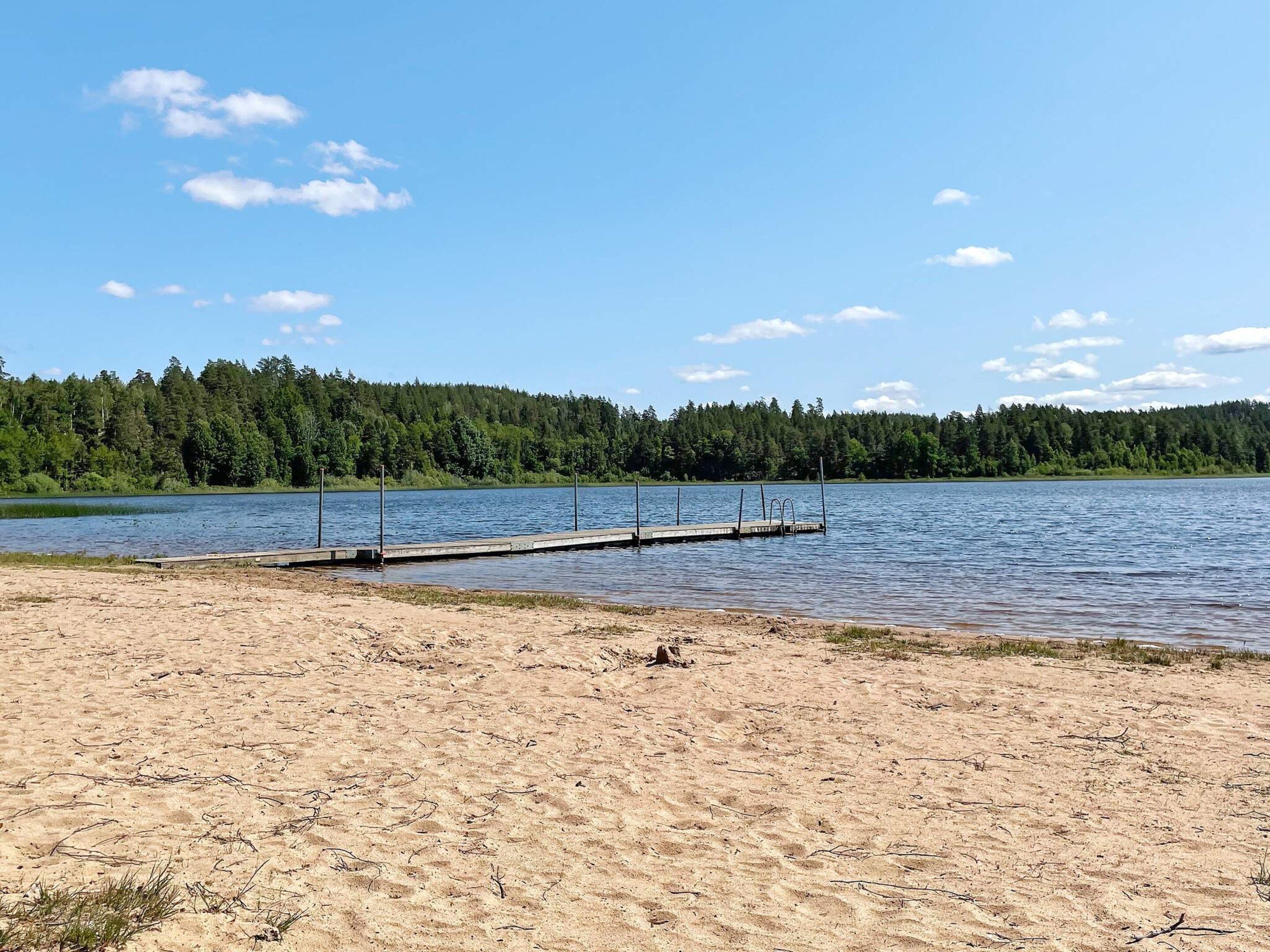 Una playa arenosa con un muelle de madera que se adentra en un lago tranquilo, rodeada de un bosque frondoso.