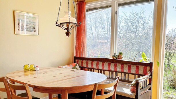 A dining area with a round wooden table, four chairs, a bench with a patterned cushion, a hanging light fixture, and a window with a view of trees and a cactus. - 6 Person Holiday Home in Borgholm (Borgholm)