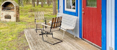 A wooden deck with a bench and a table, a stone fireplace, and a building with a red door and blue trim.