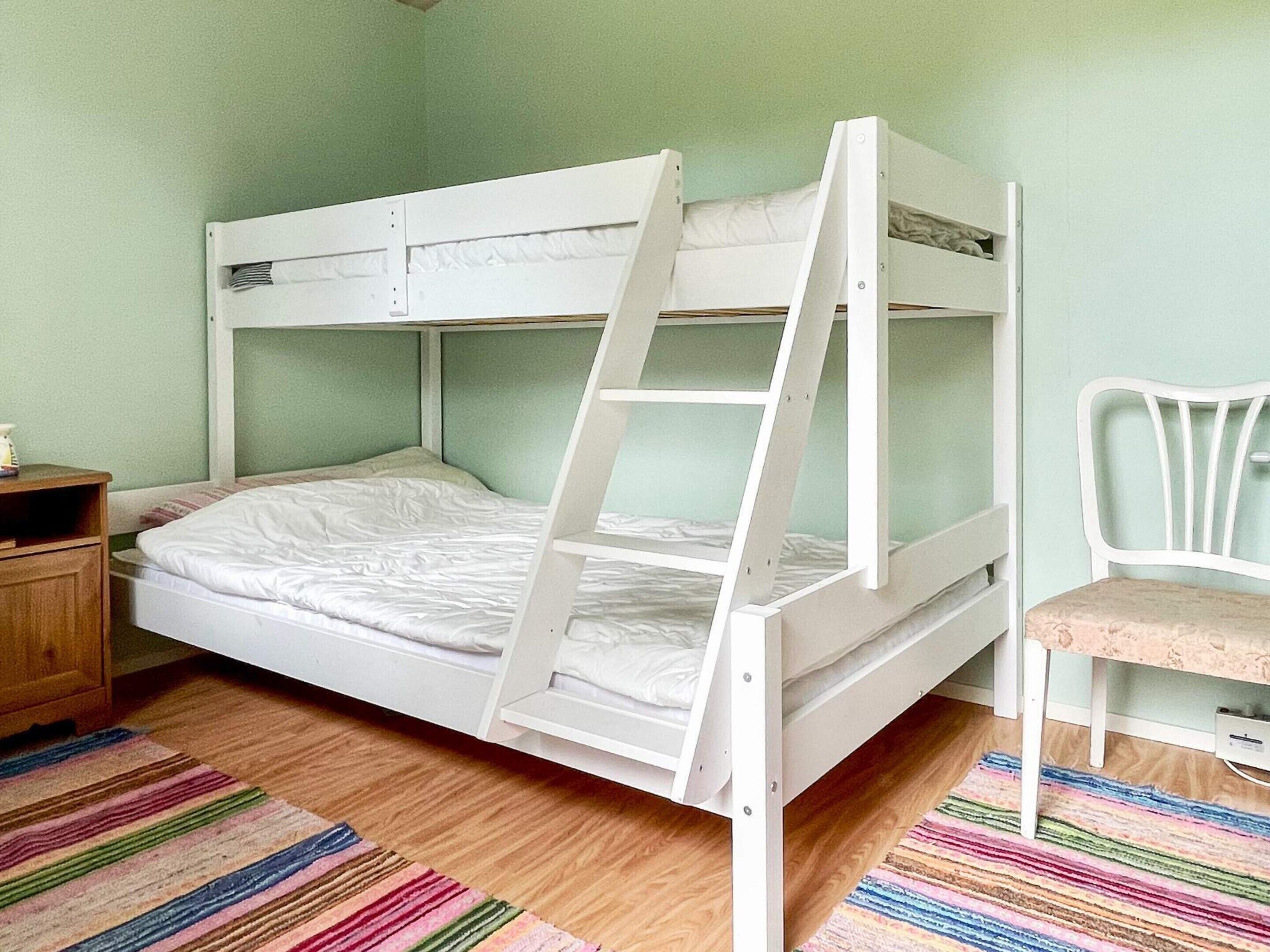 A bunk bed with a ladder, a wooden nightstand, a chair, and a striped rug in a room with light green walls.