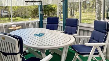 A covered outdoor seating area with a round table and chairs, a decorative hanging light fixture, and a view of greenery outside.