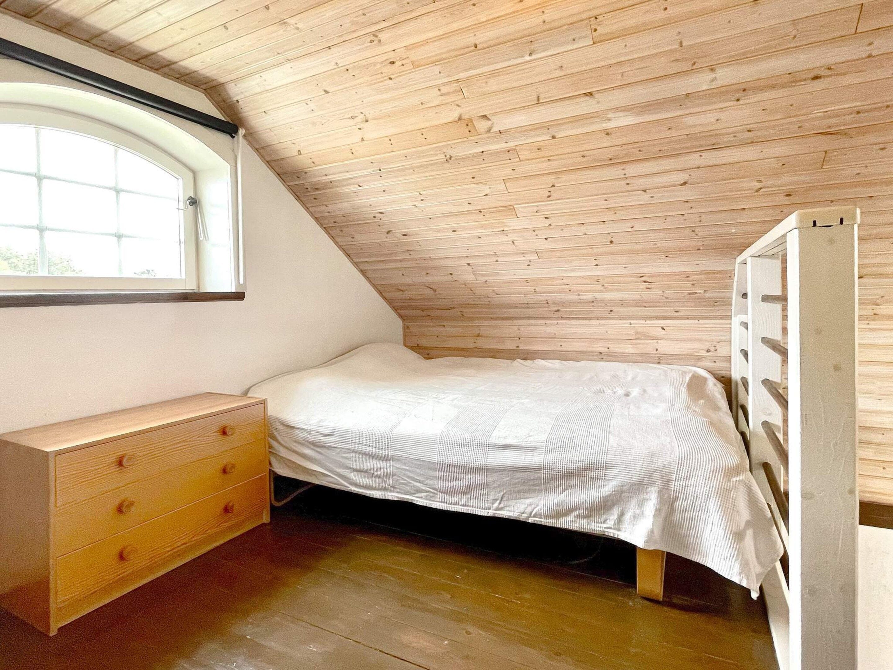 A small, single-bed room with a sloped ceiling, a wooden dresser, and a window.