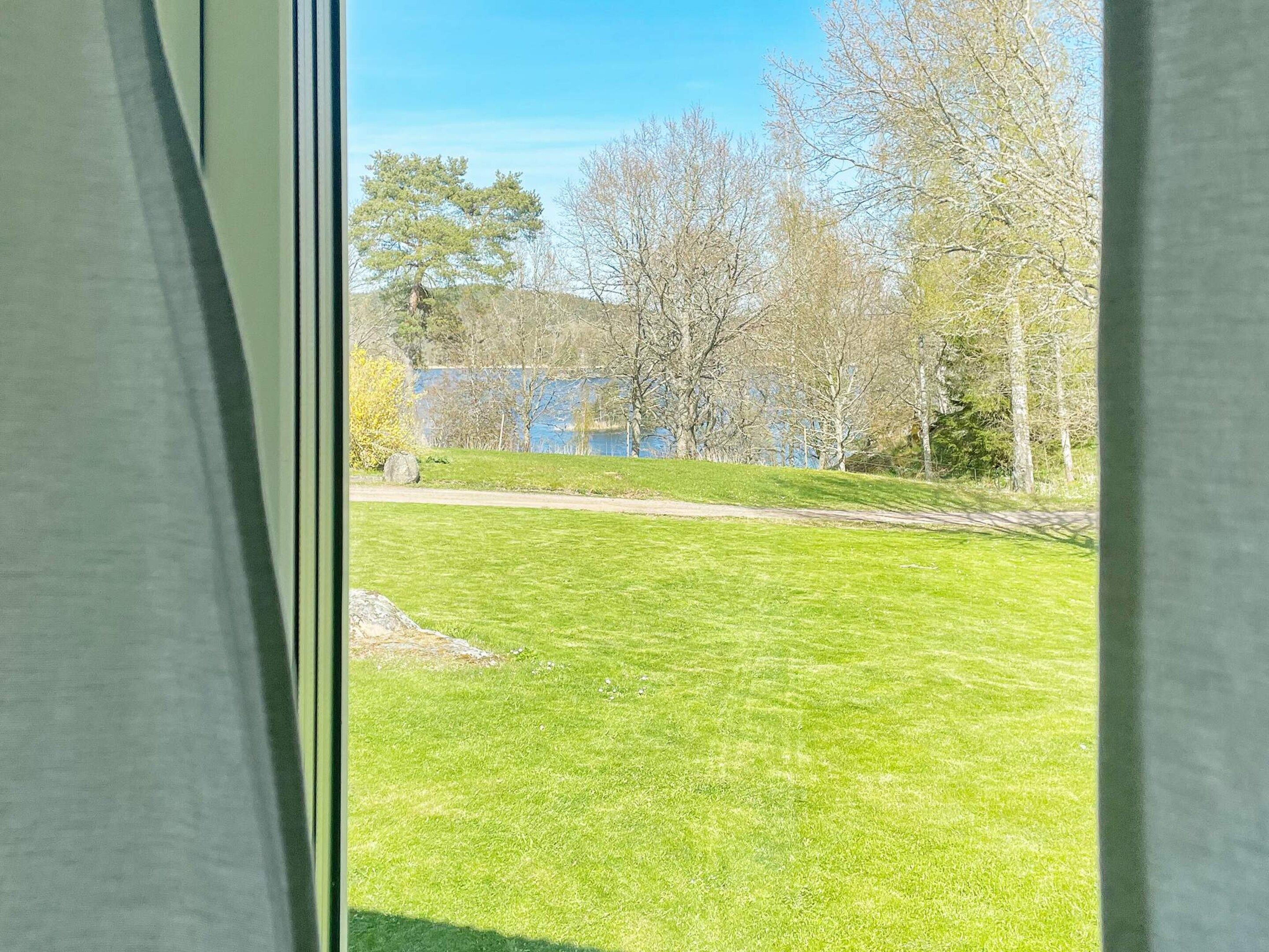 En vy genom ett fönster ut mot en gräsklädd gård med träd och en vattenyta i bakgrunden.