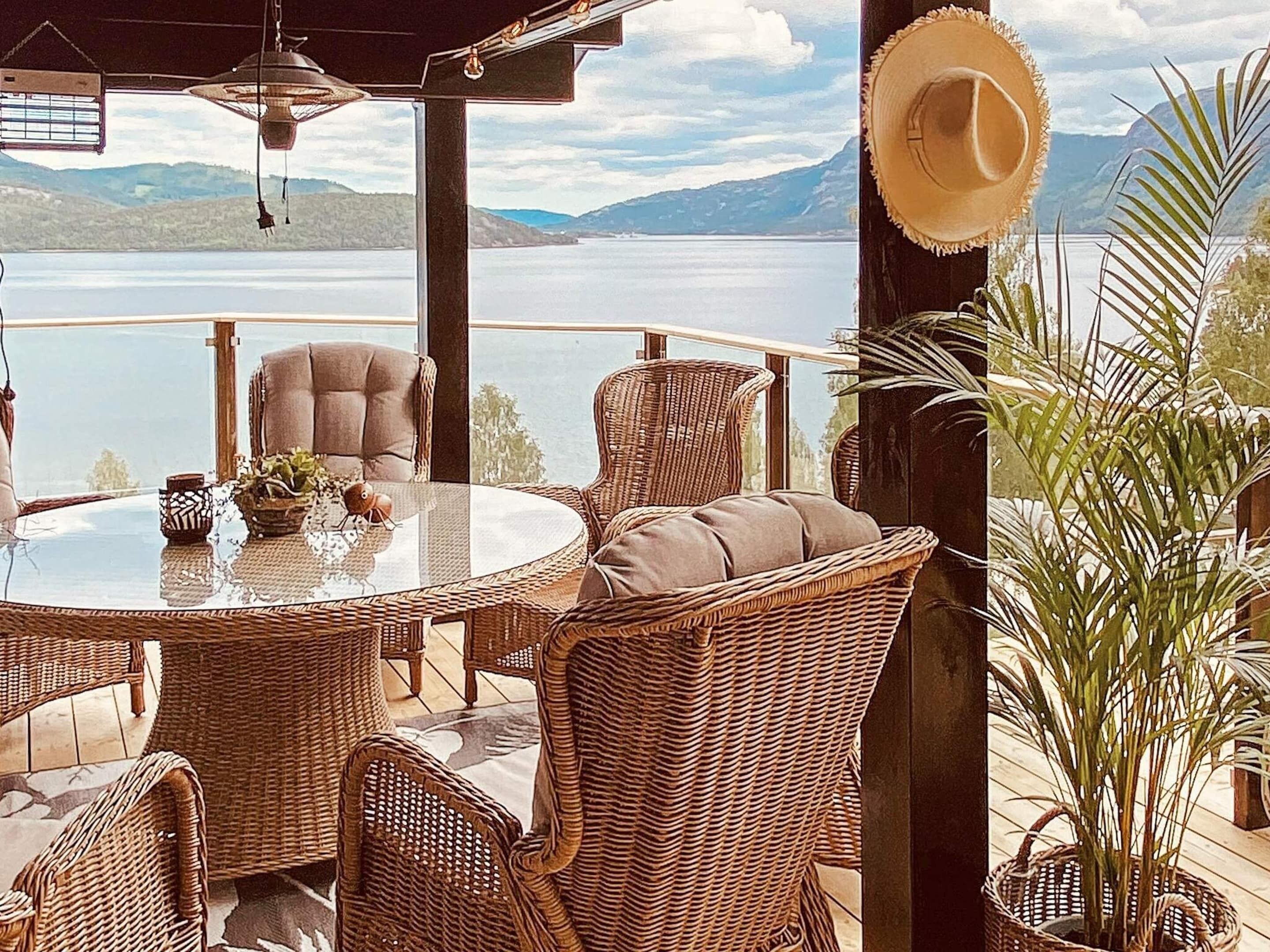 A wicker dining set with a glass table and chairs, a hat on the wall, and a view of a lake and mountains.