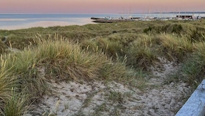 A beach with grassy dunes, a wooden boardwalk, and a marina with boats in the background. - 6 Person Holiday Home in Albaek (Aalbaek)