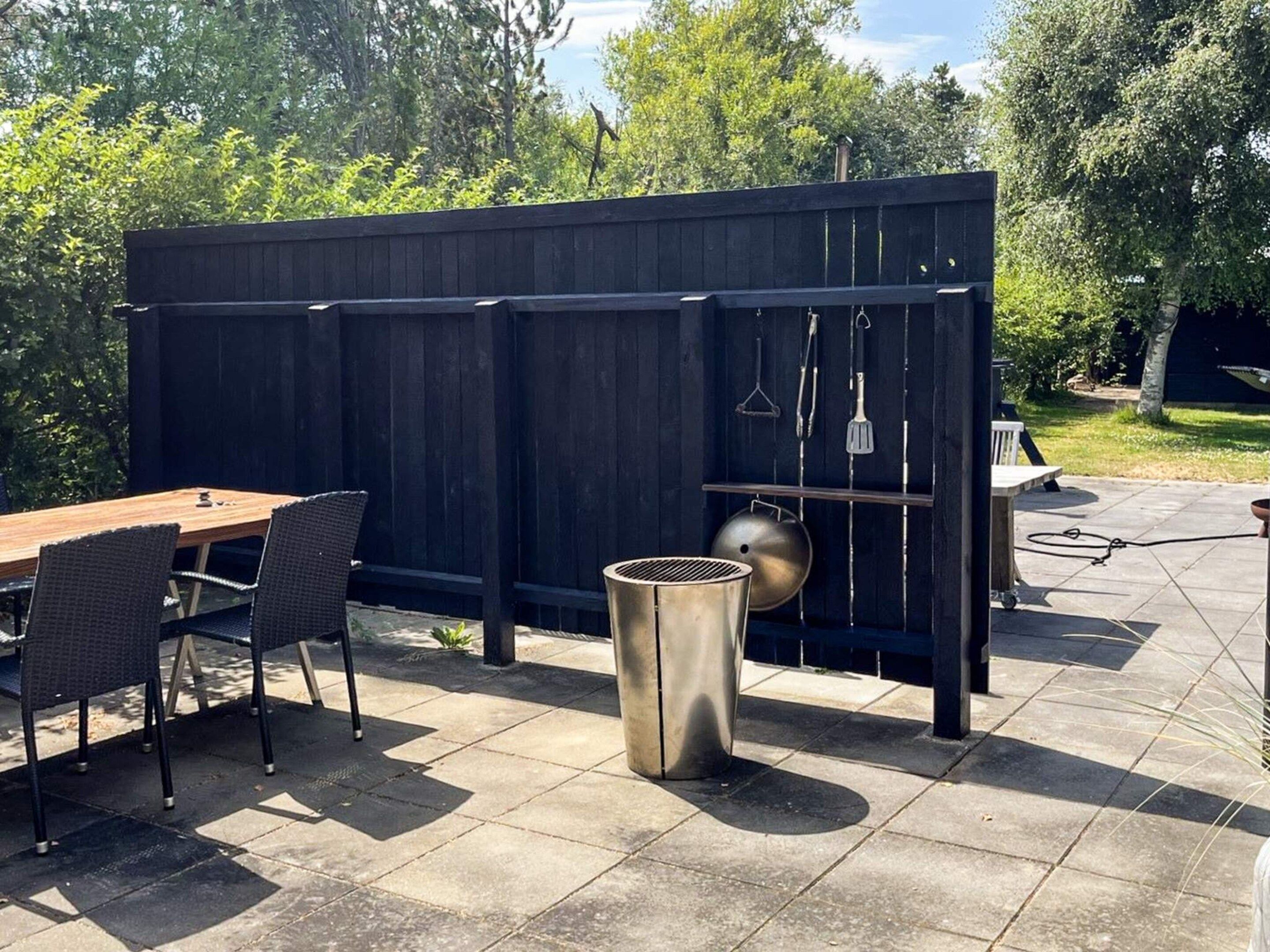 An outdoor patio with a wooden table and chairs, a metal grill, and a trash can.