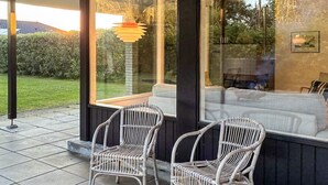 A modern outdoor seating area with two wicker chairs, a glass partition, and a view of a garden. - 6 Person Holiday Home in Albaek (Aalbaek)