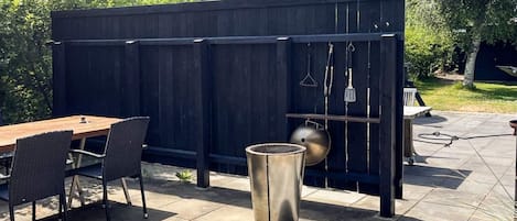 An outdoor patio with a wooden table and chairs, a metal grill, and a hanging utensil rack.