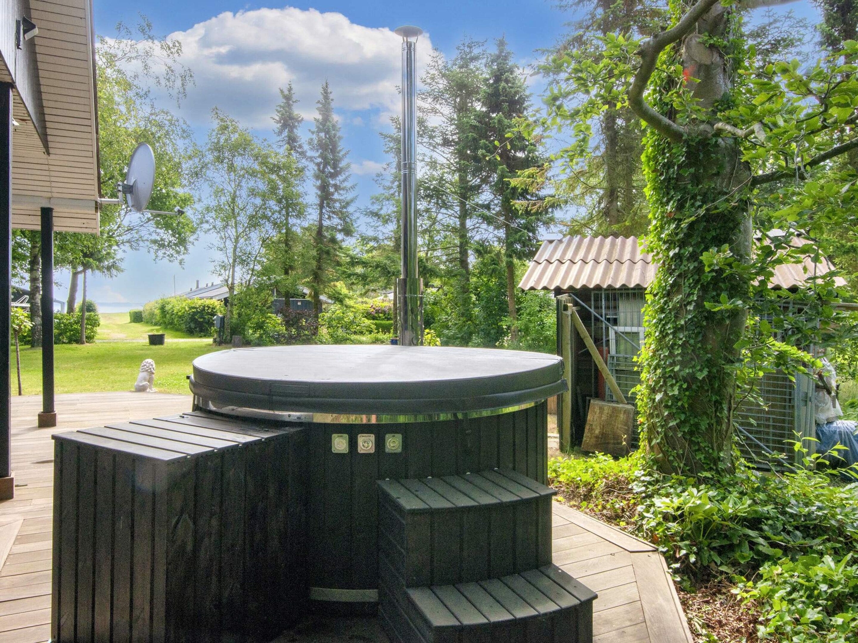 A hot tub with a wooden deck, surrounded by greenery and a view of the sky.