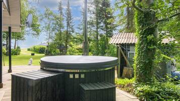 A hot tub with a wooden deck, surrounded by greenery and a view of the sky.
